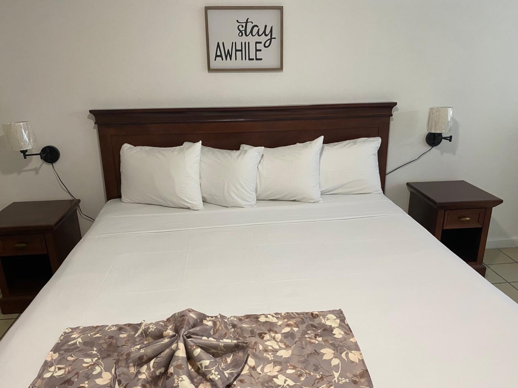 A new king room with a dark wood headboard, white linens and a “stay awhile” sign above the bed